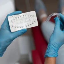 choosing-invisible-braces-orthodontist Close-up of hands in blue medical gloves presenting a clear plastic aligner alongside a card of metal brace brackets.
